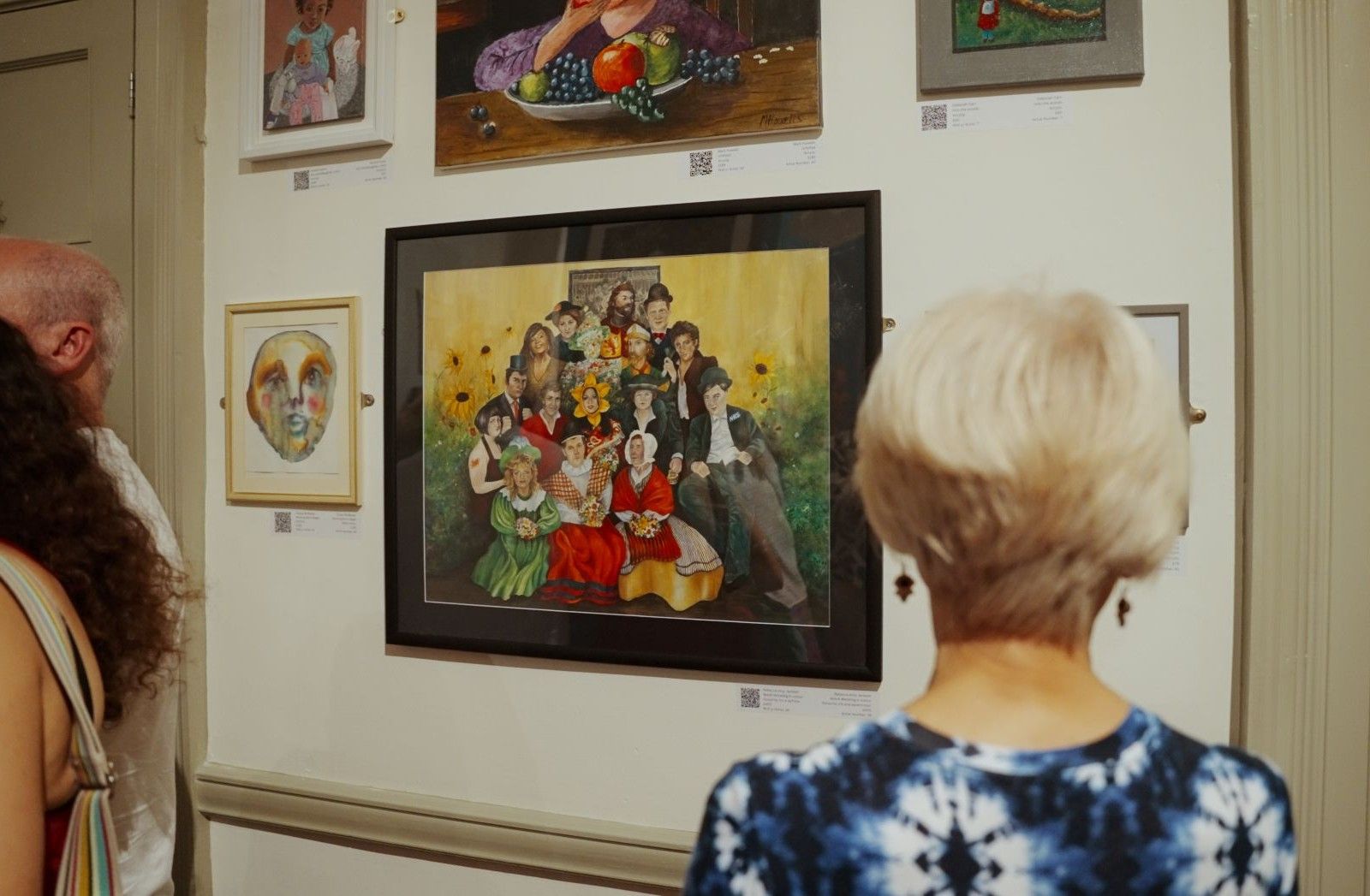 The back of a woman with short light coloured hair and a blue floral top who is looking at an artwork of a group of people..jpg