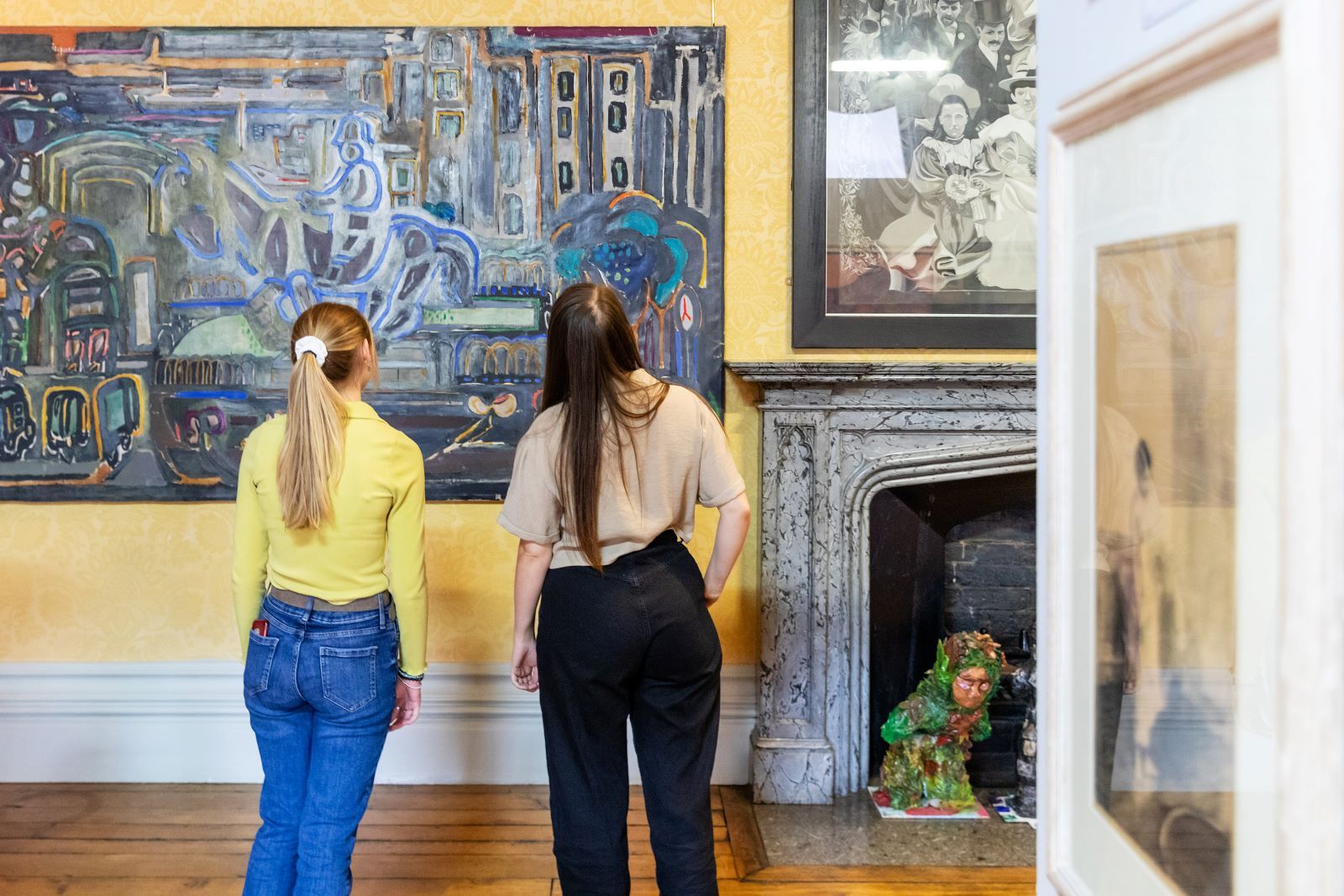 Woman with blonde ponytail and yellow top stood next to woman in beige top with brown hair looking at artwork on wall..jpg Woman with blonde ponytail and yellow top stood next to woman in beige top with brown hair looking at artwork on wall..jpg