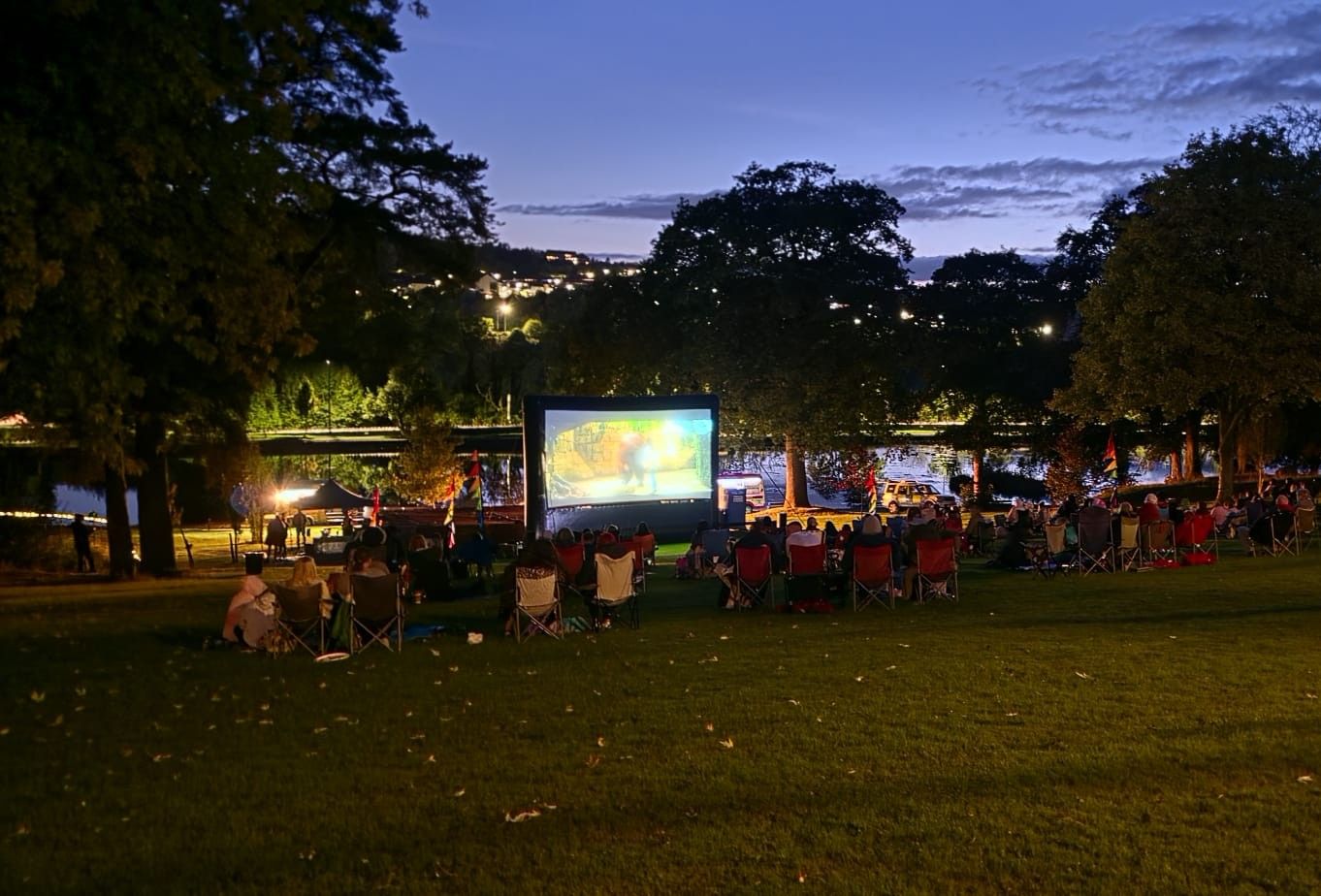 A crowd of people sat in chairs outside in front of a large screen..jpeg